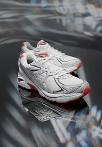 Bílé sportovní boty s červenými akcenty, síťovaný svršek a reflexní detaily. Zaoblená špička a polstrovaná podrážka dokonale integrují designové prvky.