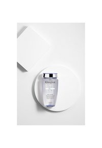 Flacon transparent de shampooing Kérastase Blond Absolu Bain Lumière sur une plateforme ronde blanche avec un carré blanc en arrière-plan.
