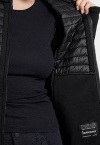 O persoană îmbrăcată cu un tricou negru cu striații dezvăluie interiorul matlasat al unei jachete negre deschise, cu eticheta de izolație Thermore de la Didriksons.
