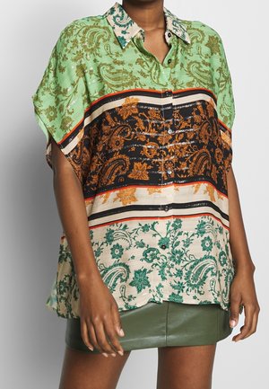 Mujer que lleva una camisa holgada de manga corta con motivos florales paisley en verde, naranja, negro y crema, y una minifalda verde de cuero.