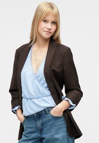 Brązowa marynarka na jasnobłękitnej bluzce z koronkowym wykończeniem, zestawiona z dżinsami z wysokim stanem. Marynarka ma podwinięte rękawy i dopasowany krój.