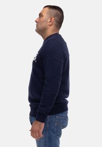 Maglione lavorato a maglia di colore blu navy con collo rotondo, polsini e orlo a coste. Indossato con jeans azzurri, visto di lato.