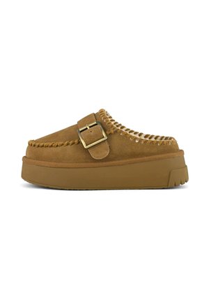 Zapato de ante marrón tipo slip-on con suela gruesa, costuras decorativas y una gran hebilla de latón en la correa lateral.