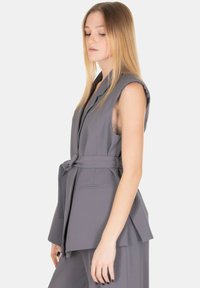 Blazer grigio senza maniche in tessuto liscio, con design strutturato, cintura in vita e tasche laterali, dal look pulito e minimalista.