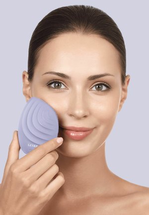 Femme à la peau claire utilisant une brosse nettoyante visage à la lavande sur sa joue droite sur un fond violet clair.