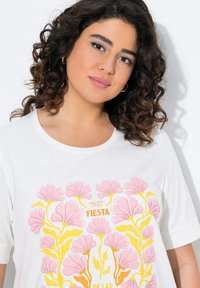 T-shirt blanc arborant un design floral avec des fleurs roses et jaunes, et le texte "1987 FIESTA DE LAS" dans des couleurs vives. Texture lisse.