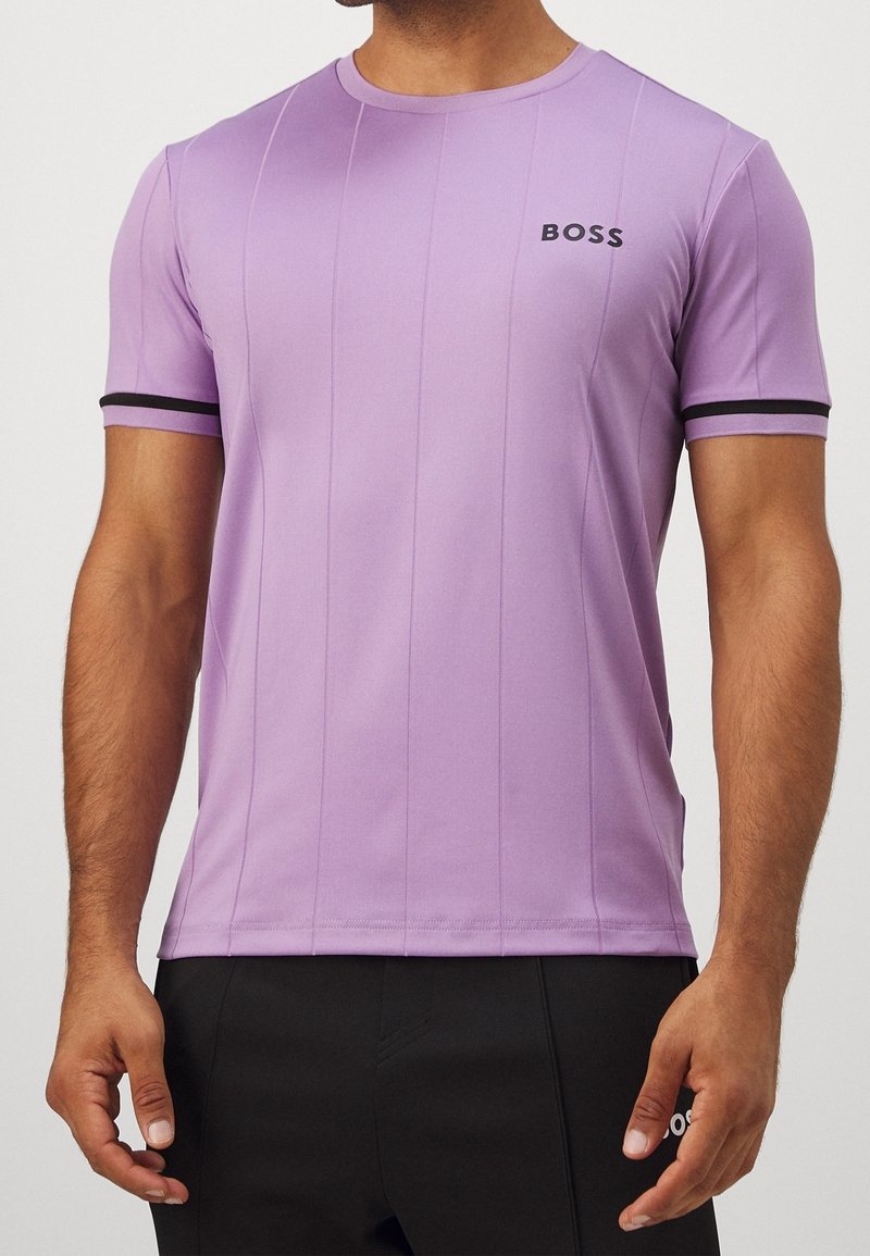 Homme portant un t-shirt ajusté lavande avec des bordures noires aux manches et le logo "BOSS" sur la poitrine, associé à un pantalon noir.