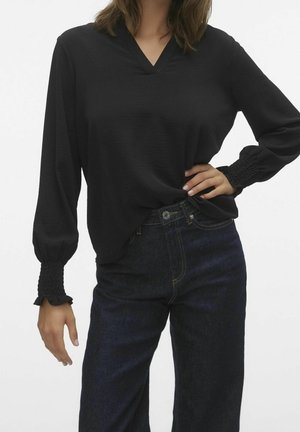 Frau trägt schwarze Bluse mit langen Ärmeln und gerafften Manschetten sowie dunkelblaue High-Waist-Jeans, Hand auf der Hüfte, weißer Hintergrund.