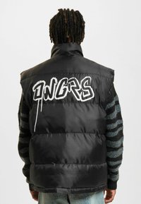Sort puffer vest med høj krave, prydet med hvid graffiti-lignende tekst på ryggen. Materialet fremstår skinnende og fyldigt.