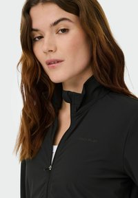 Giacca sportiva nera con collo alto e cerniera frontale. Presenta una texture liscia e un logo in lettere scure sul petto.