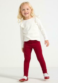 Hvid langærmet skjorte med flæsedetaljer, kombineret med røde leggings. Barnet har røde sko på og blondt hår sat med en hårnål.