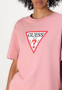 Šviesiai rožinė medvilninė T-marškinė su apvaliu kaklu. Pabrėžtas raudonas ir juodas trikampio logotipas su užrašu "GUESS U.S.A." ir klausimo ženklo dizainu.