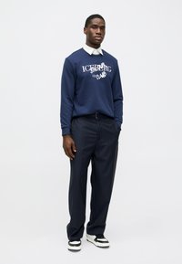 Μπλε φούτερ με λευκό κείμενο, συνδυασμένο με παντελόνια με λεπτές ρίγες. Τα αθλητικά παπούτσια έχουν μαύρο και λευκό σχέδιο. Ορατός απλός γιακάς.