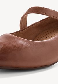 Ballerines en cuir marron avec bout arrondi, semelle intérieure rembourrée et fine sangle traversant le dessus pour un ajustement sécurisé. Texture lisse.
