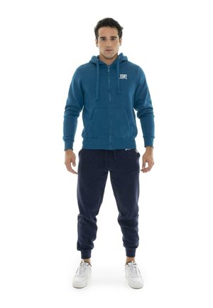 Un hombre luce una sudadera con capucha verde azulado y cremallera, pantalones de chándal azul marino y zapatillas blancas sobre un fondo blanco liso.