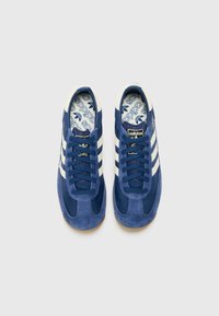 Blue suede sneakers with cream stripes, round toe, flat laces, and a textured insole featuring the adidas logo. Rubber sole for grip.