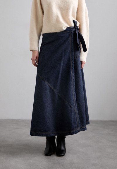 Falda envolvente de mezclilla en azul oscuro con cierre de lazo, que presenta costuras diagonales y costuras visibles. Combinada con un suéter ligero y texturizado y botas negras hasta el tobillo.