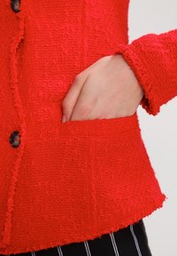Veste rouge texturée avec une fermeture à bouton et une poche avant. Le tissu a une apparence floue et tissée.
