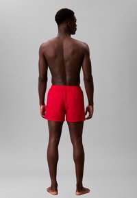 Pantaloni da bagno rossi con cintura elastica. Tessuto leggero. Il modello sta in piedi scalzo con una corporatura muscolosa, visto di spalle.