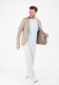 Mann, der in einem beigen Blazer über einem hellblauen T-Shirt, weißen Hosen und weißen Sneakers auf einem schlichten weißen Hintergrund geht.