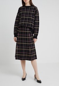 Jersey negro de punto con patrón de cuadros en rosa y dorado, combinado con una falda a juego de cuadros hasta la rodilla. Zapatos negros de tacón completan el conjunto.