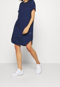 Donkerblauwe jurk met korte mouwen en een ronde halslijn, zijzakken en een gebogen zoom. Gecombineerd met witte sneakers voor een casual stijl.