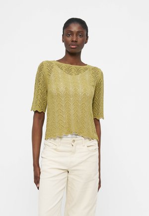 Vrouw draagt een lichtgroen, gebreid topje met korte mouwen en geschulpte randen, gecombineerd met ecru hoge taillebroek, staand tegen een effen witte achtergrond.