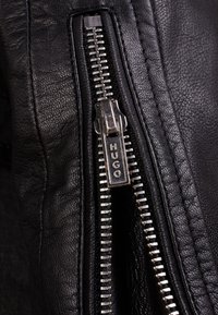 Schwarzes Leder mit Reißverschlussdetails, versehen mit einem silberfarbenen Ziehgriff, auf dem "HUGO" eingraviert ist, neben sichtbaren Nähten auf dem Stoff.