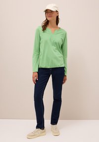 Frau in einem hellgrünen Langarmshirt, dunklen Jeans, weißen Turnschuhen und einer weißen Baseballmütze, die vor einem beigen Hintergrund steht.