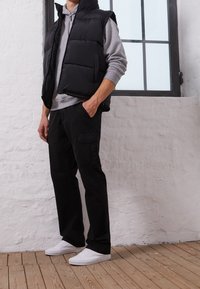 Chaleco acolchado negro sobre una sudadera con capucha gris, combinado con pantalones cargo negros y zapatillas blancas. El entorno tiene un suelo de madera clara y una ventana.