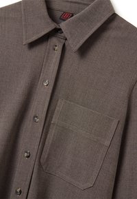 Camicia button-up di colore marrone con colletto a punta e tasca sul petto, in cui vengono mostrati dettagli ravvicinati del tessuto e dei bottoni.