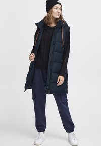 Mörkblå puffer väst med hög krage och orange dragband, kombinerad med en svart kabelstickad tröja och mörkblå joggers. Vita sneakers.