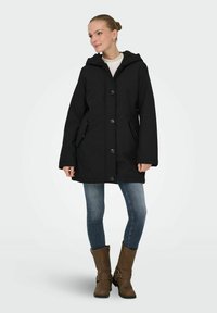 Crna kaput s kapuljačom, zatvoren dugmadima, s dva bočna džepa i opuštenim krojem. Uparena s plavim farmerkama i smešnim čizmama do sredine lista.