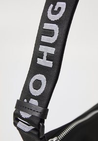 La tracolla nera presenta un audace testo bianco che dice "NO HUG." La tracolla è realizzata in tessuto testurizzato con un elegante accento in pelle nera liscia.