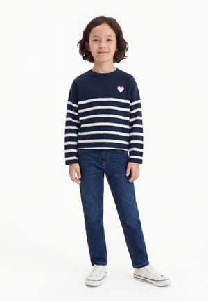 Enfant debout portant un t-shirt manches longues rayé bleu marine et blanc avec un cœur rose, un jean bleu et des baskets blanches sur fond blanc.