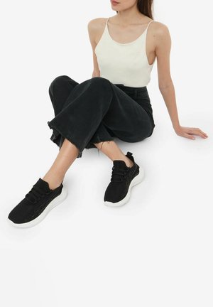 Sneaker da atletica neri con una tomaia texturizzata, lacci elastici e suola in gomma bianca, abbinati a una canotta crema e jeans neri.