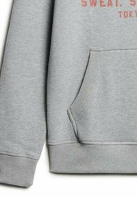Grå sweatshirt med kengurulomme, ribbede manchetter og kant; blødt stof med rødt trykt tekst, der står "SWEAT: TOKYO."