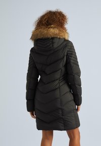 Sieviete ar cirtainiem matiem, ģērbta melnā pamatīgā ziemas mētelī ar kažokādas oderi, redzama no mugurpuses.