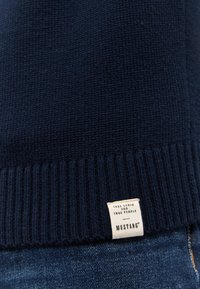 Sötétkék kötött pulóver, bordás aljjal, látható márkacímkével, amely a "True Denim for True People" és a "MUSTANG" feliratot tartalmazza.