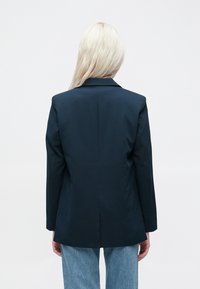 Jūrinis bleizeris su struktūrizuotu dizainu, išpjautomis atvartais ir vienu nugariniu plyšiu. Lygus audinys, pritaikytas fasonas, derinamas su šviesiai mėlynomis džinsomis.