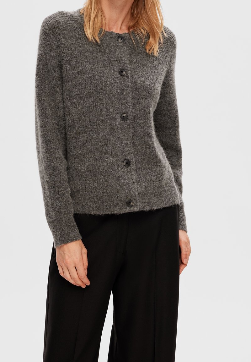 Cárdigan de punto gris con cuello redondo y seis botones negros, con una textura suave y mangas largas. Combinado con pantalones negros.