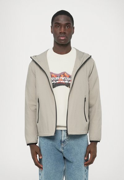 Hellgraue Jacke mit Reißverschluss aus strukturiertem Stoff, verstellbarer Kapuze und schwarzen Akzenten. Getragen über einem weißen Sweatshirt mit Grafik und blauen Jeans.