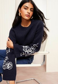 Pulover navy cu broderie florală albă pe mâneci, material tricotat texturat și mansete cu elastic. Asortat cu blugi blue.