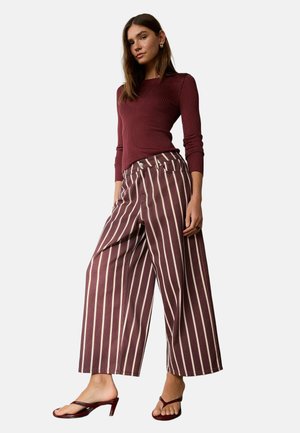 Mujer con top ajustado de canalé burdeos, pantalones burdeos de pierna ancha con rayas verticales blancas y sandalias burdeos de tacón, de pie sobre fondo blanco.