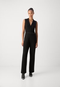 Combinaison noire avec un buste ajusté, décolleté en V et accents cloutés aux épaules et à la taille. Jambes larges et texture lisse, associée à des bottes noires à talons.