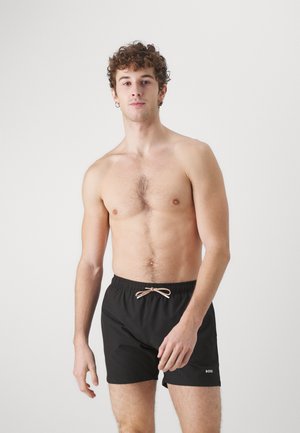 Fiatal férfi göndör hajjal, ing nélkül, fekete zsinóros rövidnadrágot visel, melyen "BOSS" logó látható, egyszerű fehér háttér előtt áll.