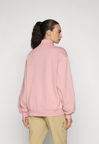Felpa rosa oversize con colletto alto e spalle cadenti, caratterizzata da una texture morbida e polsini a costine, abbinata a pantaloni cargo beige.