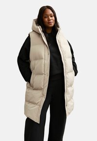 Chaleco acolchado beige con capucha, que presenta un diseño acolchado, bolsillos frontales y un ajuste relajado, que se lleva sobre una camiseta de manga larga negra y pantalones.