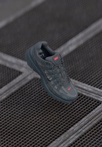 Nike sneakers in dark grey with a textured mesh upper, contrasting red accent logo, and rubber sole, featuring a grid pattern design.
