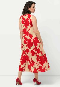 Bezramínková béžová šaty s velkým červeným květinovým vzorem; vrstvený design s volánovým lemem. Kombinováno s červenými vysokými podpatky.